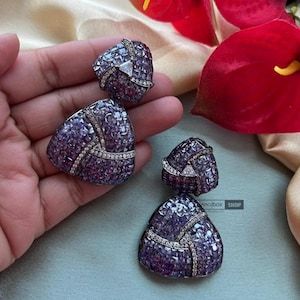 May include: A pair of dangle earrings with a triangular shape. The earrings are made of silver and are adorned with purple and clear stones.