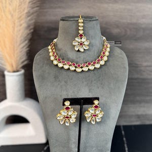 May include: A gold necklace, earrings, and a tikka with white and pink stones. The necklace has two rows of stones and the earrings and tikka have a flower design.
