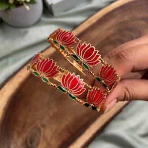 May include: Two gold-colored bangles with red lotus flower designs and green accents. The bangles are displayed on a wooden surface, showcasing the intricate floral patterns and vibrant colors.