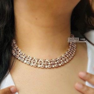 Peut inclure: Un collier ras de cou en or avec trois rangées de pierres de zircone cubique transparentes. Le collier est superposé et a un look délicat et féminin.