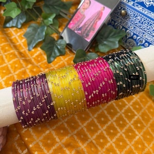 May include: A collection of colorful glass bangles in shades of purple, yellow, pink, and green, adorned with small gold-colored accents. The bangles are displayed on a white cylindrical support, set against a patterned yellow fabric. A smartphone displays an image of a woman.