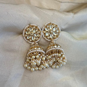 以下が含まれることがあります： 金色のジュムカピアス。各イヤリングは、透明な石を使った花の模様で、小さな白いビーズで囲まれています。ベル型の底には、ぶら下がっているイミテーションパールが飾られています。