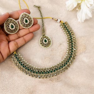 May include: A gold necklace with a teardrop design, featuring green and white stones. The necklace is paired with matching earrings and a maang tikka.