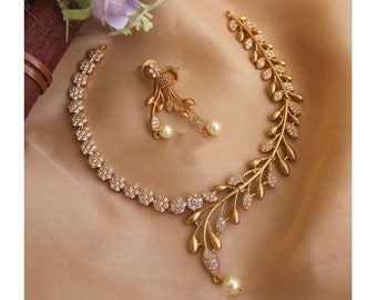May include: A gold-coloured jewellery set comprising a necklace and earrings. The necklace features a floral design with clear stones and a leaf-like branch with clear stones. The earrings have a leaf design with a pearl. The set is on a beige surface.