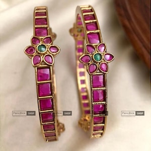 Może przedstawiać: Dwie bransoletki w kolorze złota z kwadratowymi, magentowymi kamieniami. Każda bransoletka ma kwiatowy wzór z magentowymi płatkami i zielonym kamieniem centralnym. Bransoletki są prezentowane na białej powierzchni.