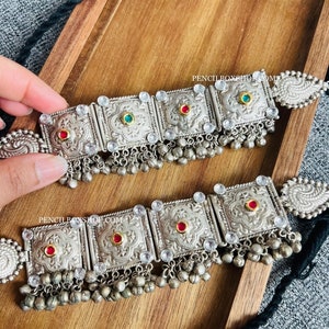 May include: A silver choker necklace with red and green stones, intricate detailing, and dangling silver beads.
