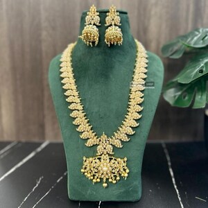 May include: A gold necklace with a pendant shaped like a flower with pearl accents. The necklace is made of gold-plated metal and has a traditional Indian design. The necklace is displayed on a green velvet stand. The necklace comes with a matching pair of earrings.