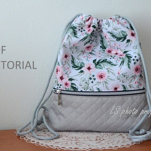 Op de afbeelding: Een grijze rugzak met trekkoord en een bloemenprint en een ritsvak. De rugzak heeft een grijs trekkoord en de tekst "LS photo props" is onderaan gedrukt. De tekst "PDF TUTORIAL" is op de muur achter de rugzak gedrukt.