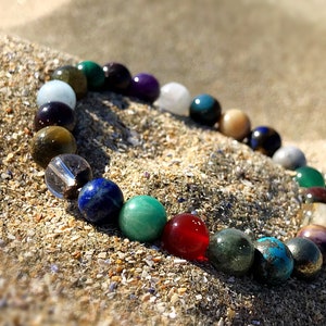 Op de afbeelding: Een kleurrijke kralen armband met verschillende edelstenen, waaronder groen, blauw, rood en bruin, op een zanderige achtergrond.