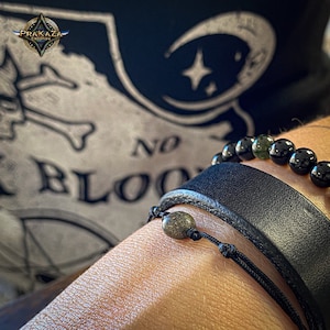 Peut inclure: Gros plan sur le poignet d'une personne portant deux bracelets. Le bracelet du haut est composé de perles noires avec des accents verts. Le bracelet du bas est un bracelet en cuir noir avec une seule perle de pierre verte.