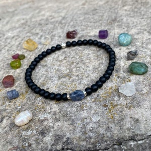 Peut inclure: Un bracelet en perles noires avec un accent argenté et une pierre précieuse bleue. Le bracelet est entouré de diverses petites pierres précieuses de différentes couleurs.