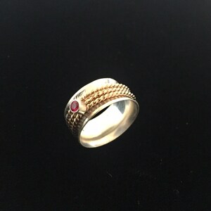 May include: A silver ring with a gold-colored rope design and a small, round, red gemstone. The ring has a polished finish and a classic, elegant style. The gemstone is set in a bezel setting.