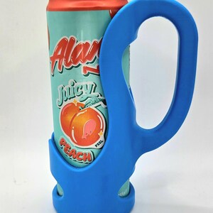 Op de afbeelding: Een blauwe blikjeshouder met een handvat, met een blikje Alani Nu Juicy Peach-frisdrank. Het blikje heeft een turquoise achtergrond met perzikafbeeldingen en rode en witte tekst. De houder is gemaakt van flexibel materiaal.