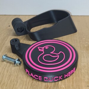 May include: A black 3D printed duck holder with a pink rubber duck outline and the text "Place Duck Here".