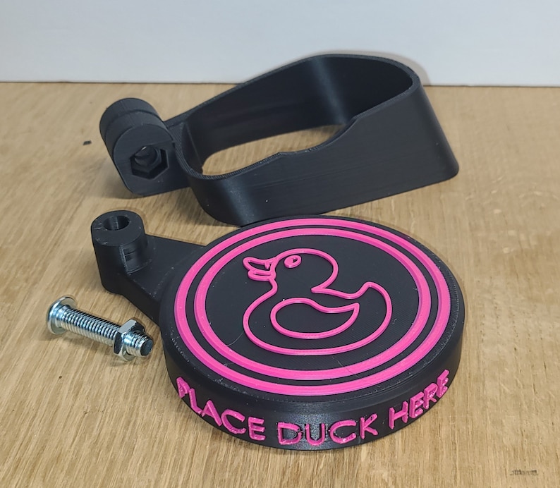 May include: A black 3D printed duck holder with a pink rubber duck outline and the text "Place Duck Here".
