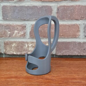 May include: A gray, 3D-printed cup holder with a unique, open design. The holder features a curved handle and a base with a circular opening. The product is made of plastic and sits on a wooden surface.