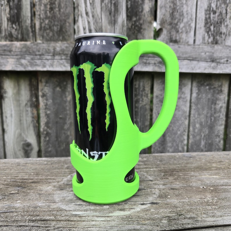 May include: A neon green can holder with a large handle, designed to fit a standard beverage can. The holder is wrapped around a black can with a green logo. The can is resting on a wooden surface.