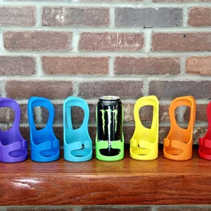 May include: A collection of colorful can holders in pink, purple, blue, teal, yellow, orange, red, and gray. A black and green Monster energy drink can is in one of the holders. The holders are on a wooden surface.