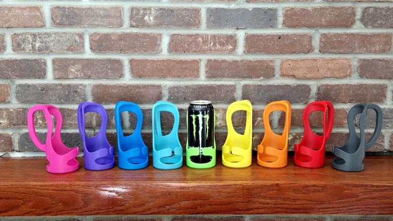 May include: A collection of colorful can holders in pink, purple, blue, teal, yellow, orange, red, and gray. A black and green Monster energy drink can is in one of the holders. The holders are on a wooden surface.
