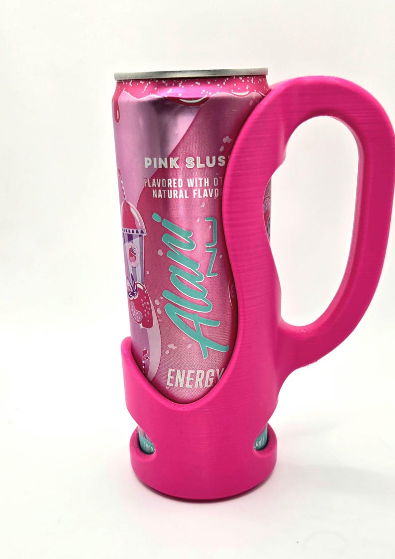 Op de afbeelding: Een roze blikje Alani Nu Energy drink in een bijpassende roze houder. Het blikje is voorzien van het opschrift "Pink Slush" en heeft een aardbei- en ijsje-afbeelding. De houder heeft een handvat.