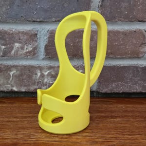May include: Bright yellow, open-design cup holder with a curved handle. The holder has a circular base and a curved, open design. The holder is made of a smooth, solid material and is designed to hold a cup or bottle.