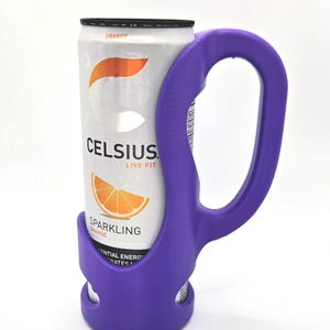 Op de afbeelding: Een paarse blikhouder met een handvat, met een blikje Celsius Sparkling Orange. Het blikje is wit met oranje afbeeldingen en tekst. De houder is gemaakt van plastic en heeft een gebogen handvat.