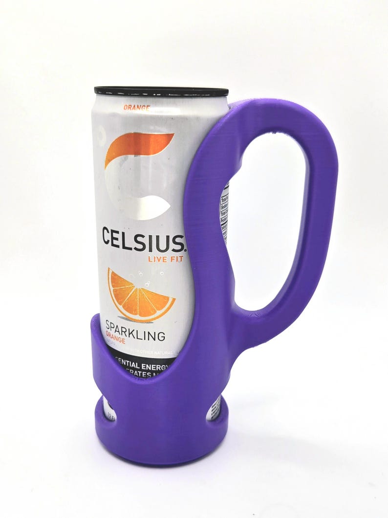 Op de afbeelding: Een paarse blikhouder met een handvat, met een blikje Celsius Sparkling Orange. Het blikje is wit met oranje afbeeldingen en tekst. De houder is gemaakt van plastic en heeft een gebogen handvat.