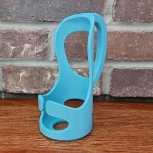 May include: A light blue, 3D-printed phone holder. The holder has a curved handle and a circular base with a hole. The design includes a slot to hold a phone. The holder is made of plastic and sits on a wooden surface.