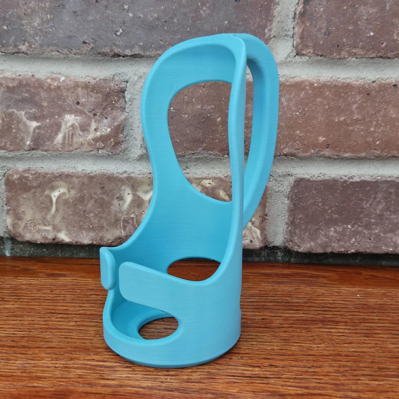 May include: A light blue, 3D-printed phone holder. The holder has a curved handle and a circular base with a hole. The design includes a slot to hold a phone. The holder is made of plastic and sits on a wooden surface.