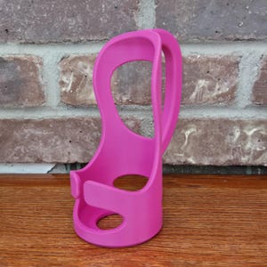 May include: A bright pink, 3D-printed cup holder. The holder has a curved handle and a circular opening at the base. The design includes a circular opening in the middle and a small clip on the side. The holder is made of plastic and sits on a wooden surface.