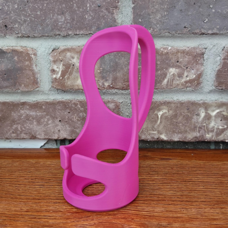 May include: A bright pink, 3D-printed cup holder. The holder has a curved handle and a circular opening at the base. The design includes a circular opening in the middle and a small clip on the side. The holder is made of plastic and sits on a wooden surface.