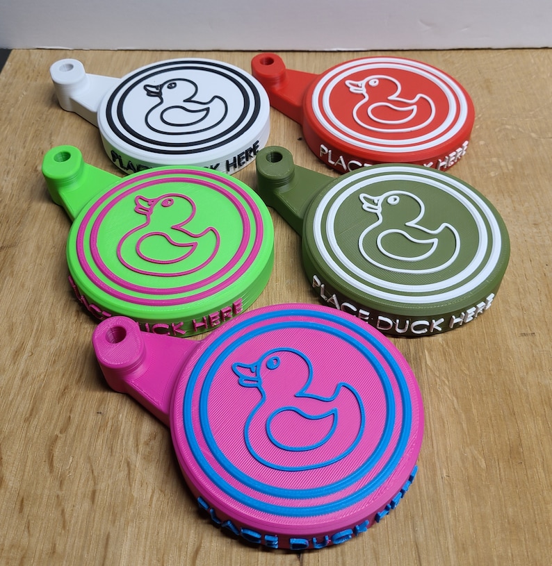 May include: Five colorful 3D printed rubber duck holders with the text "Place Duck Here". Each holder features a different color and a white rubber duck outline.