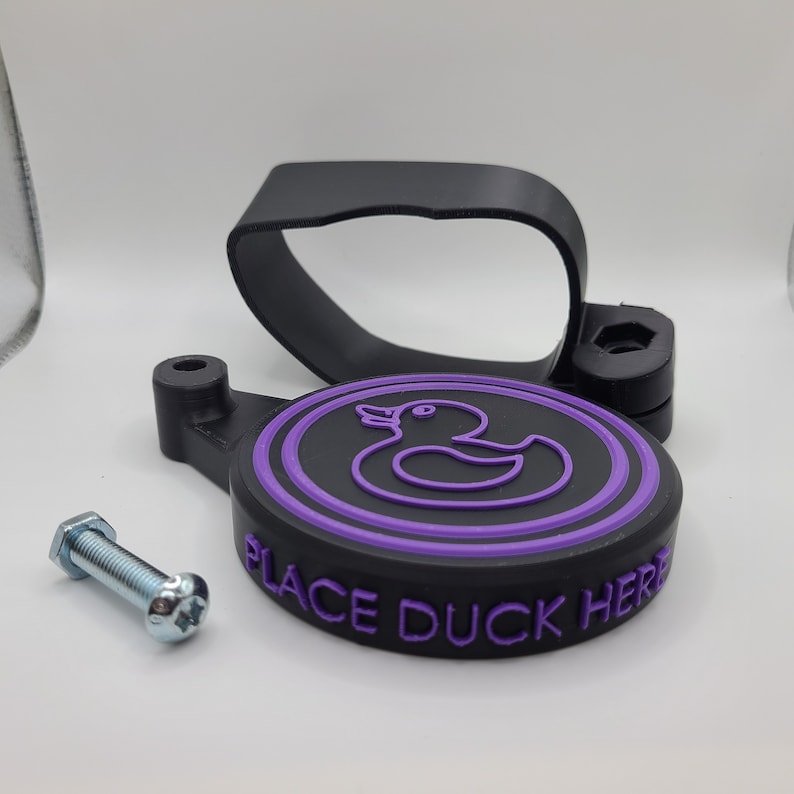 May include: A black 3D printed duck holder with a purple rubber duck outline and the text "PLACE DUCK HERE".