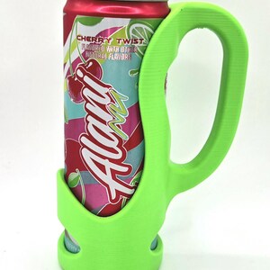 Op de afbeelding: Een felgroene blikhouder met een handvat, ontworpen om een blikje Alani Cherry Twist-frisdrank vast te houden. Het blikje is rood met een kleurrijk etiket met kersen en de merknaam. De houder biedt een stevige grip.