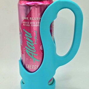 Op de afbeelding: Een roze energiedrankblikje met de tekst "Pink Slush" en "Alani Nu Energy" wordt vastgehouden door een helderblauwe blikjeshouder. De houder heeft een handvat.