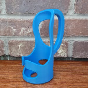 May include: A vibrant blue, 3D-printed cup holder with a unique, open design. The holder features a handle and a base with a circular opening. The product is made of plastic and is designed to hold a cup or bottle.
