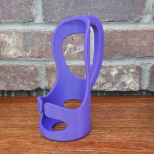 May include: A vibrant purple cup holder, designed with a curved handle and a base with a circular opening. The holder features a secure clasp and is made of a smooth, solid material. The design is modern and functional.
