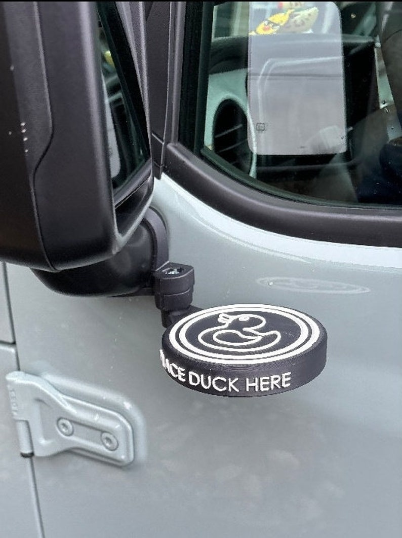 May include: A black and white rubber ducky decal attached to a car's side mirror. The decal says "Place Duck Here".