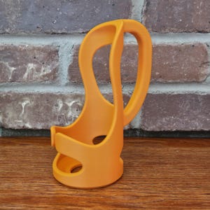 May include: Orange 3D-printed cup holder with a curved handle. The holder has a unique, open design with two circular cutouts. The handle curves upwards, providing a comfortable grip. The holder is made of a solid, smooth material.