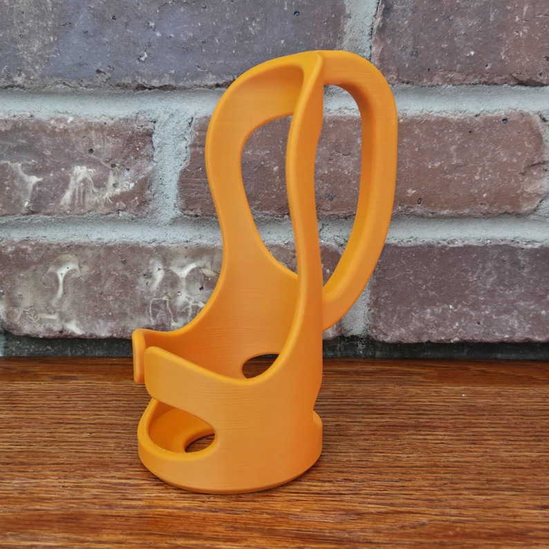 May include: Orange 3D-printed cup holder with a curved handle. The holder has a unique, open design with two circular cutouts. The handle curves upwards, providing a comfortable grip. The holder is made of a solid, smooth material.