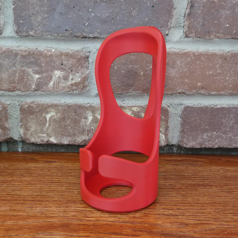 May include: A bright red, open-design holder, possibly for a beverage container, with a curved shape and a large central opening. The holder is made of a smooth material and sits on a wooden surface against a brick wall.