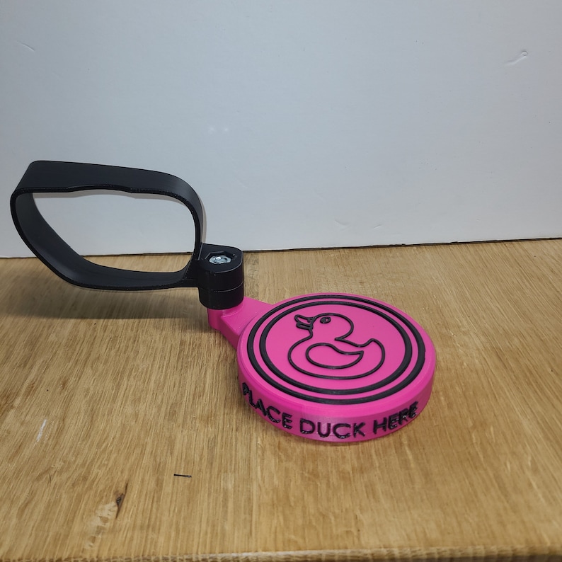 May include: A pink 3D printed duck holder with a black handle. The holder has a black outline of a duck and the words "Place Duck Here" around the bottom.