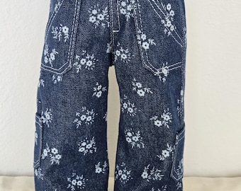 Puede incluir: Pantalones de muñeca de mezclilla azul con un estampado floral blanco. Los pantalones tienen un diseño de pierna ancha, bolsillos y costuras blancas. Los pantalones de muñeca están diseñados para adaptarse a una muñeca.