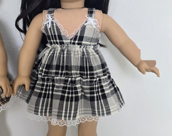 May include: A doll wearing a black, white, and gray plaid dress with white lace trim. The dress has a tiered skirt and spaghetti straps with white bows. The doll has long black hair.