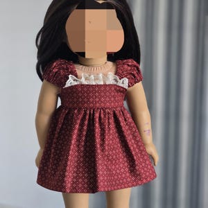 May include: A doll wearing a burgundy dress with a white dotted pattern. The dress features short, puffed sleeves and a white lace trim around the neckline. The doll has long, dark brown hair.
