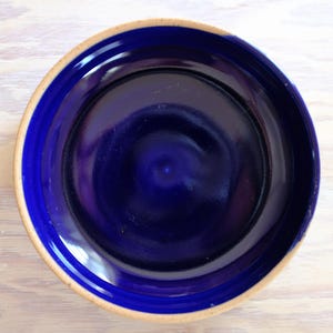 以下が含まれることがあります： 深みのある青色のセラミックプレート。幅広でわずかに盛り上がった縁が特徴です。プレートの内側は光沢のある濃い青色で、明るい無釉の縁と対照的です。明るい色の木製の表面に置かれています。
