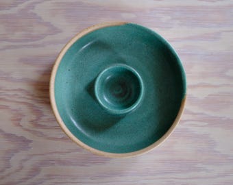 May include: A handmade teal ceramic chip and dip serving dish. The dish has a circular shape with a raised center bowl. The rim is a light tan color. The dish is designed for serving snacks and appetizers.