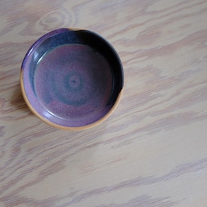 May include: A handmade ceramic bowl with a circular shape. The interior is a gradient of purple and teal, with a dark blue rim and a tan-colored edge. The bowl is set against a light wood grain background.