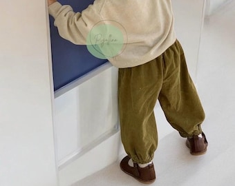 May include: A beige long-sleeved knit sweater paired with olive green corduroy trousers featuring elasticized cuffs at the ankles. The outfit is completed with dark brown leather Mary Jane shoes. A circular logo with the text Pigaffino is visible.