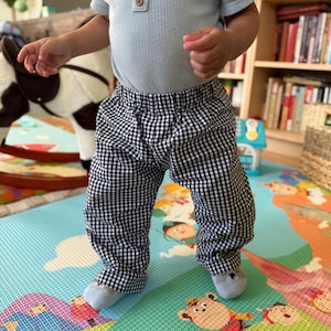 May include: A toddler wearing a light blue shirt and black and white checkered pants. The pants have an elastic waistband.